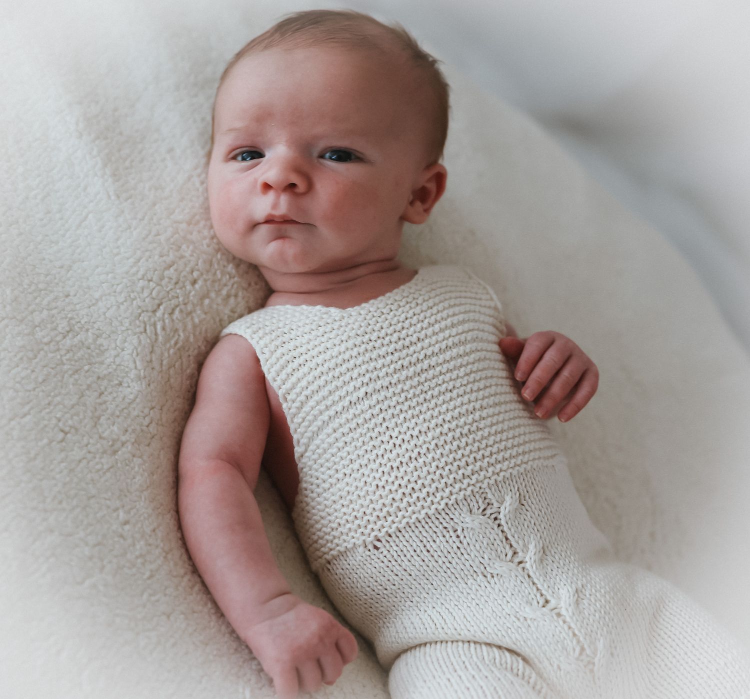Baby wearing white knitted romper lying on soft white blanket.