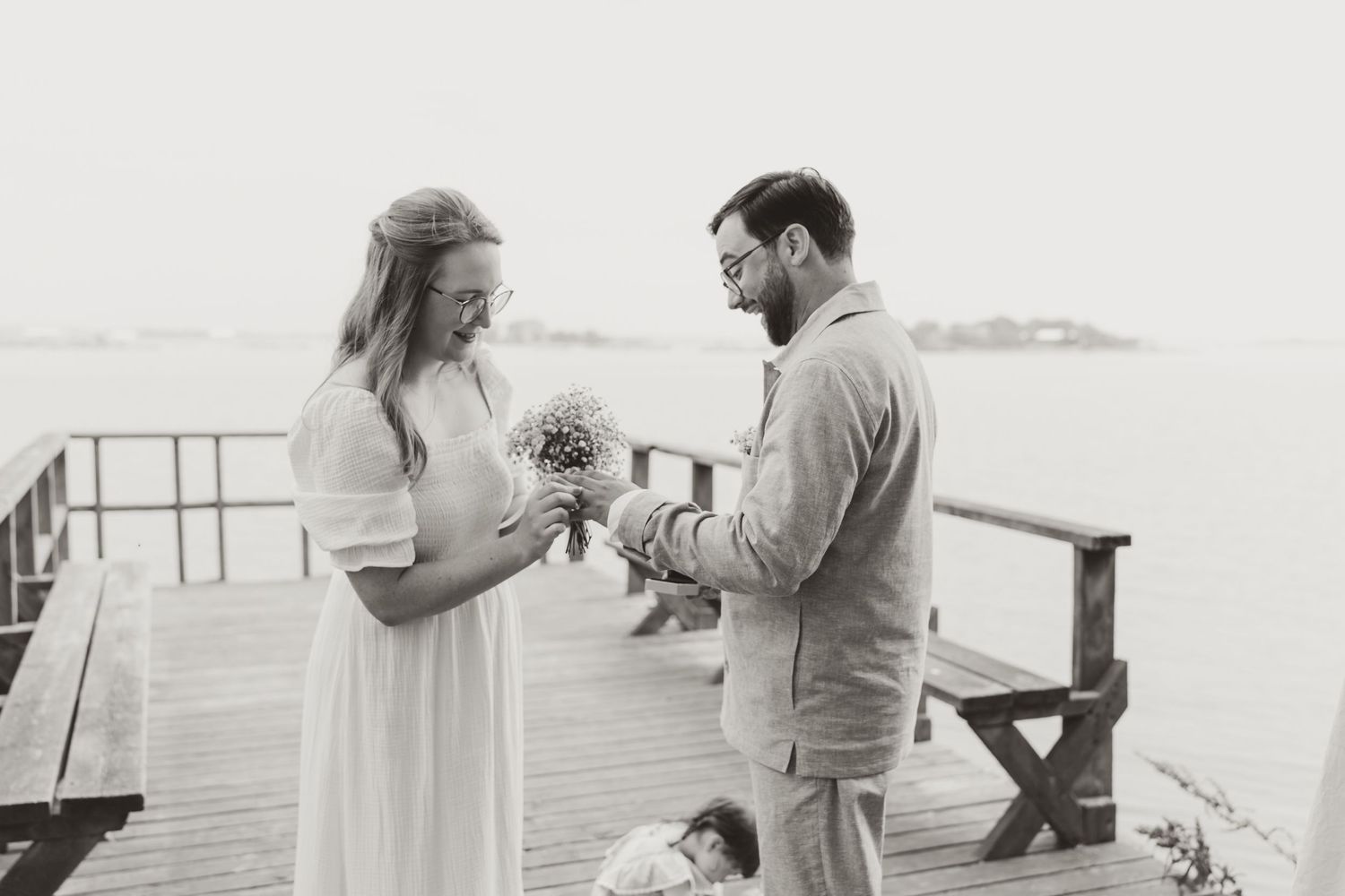 A black and white wedding moment captured on a wooden pier by the ocean with a romantic exchange of flowers.