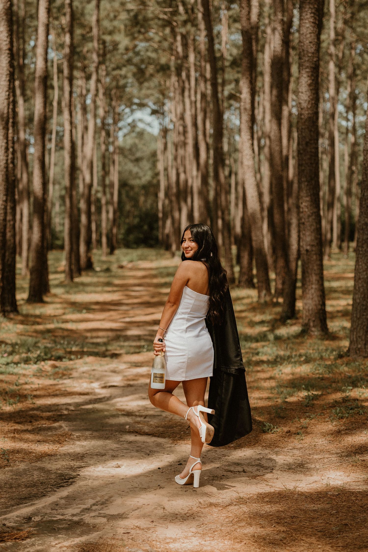 Woman in white dress standing on forest path among tall pine trees on a sunny day.