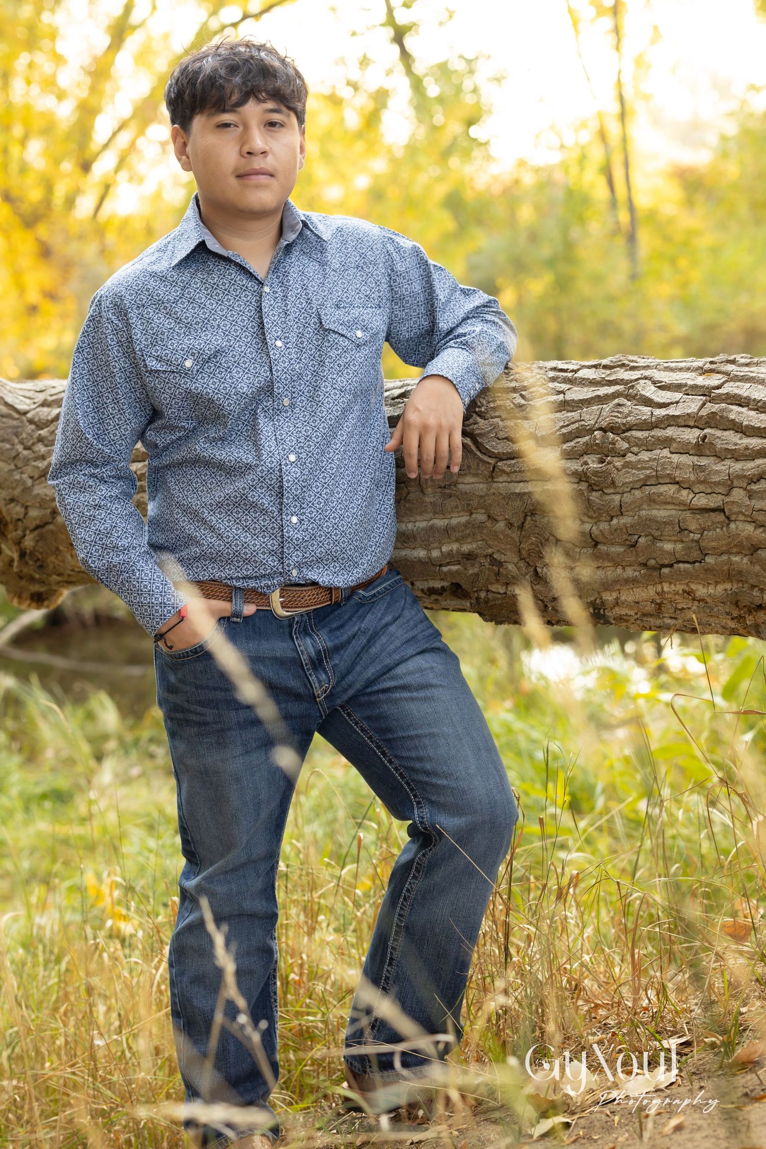 A person in a polka dot button-down shirt and jeans poses casually against a fallen log in an autumn field.