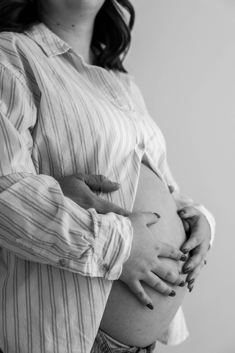 Black and white close-up of a pregnant belly being cradled by hands in a striped button-down shirt.