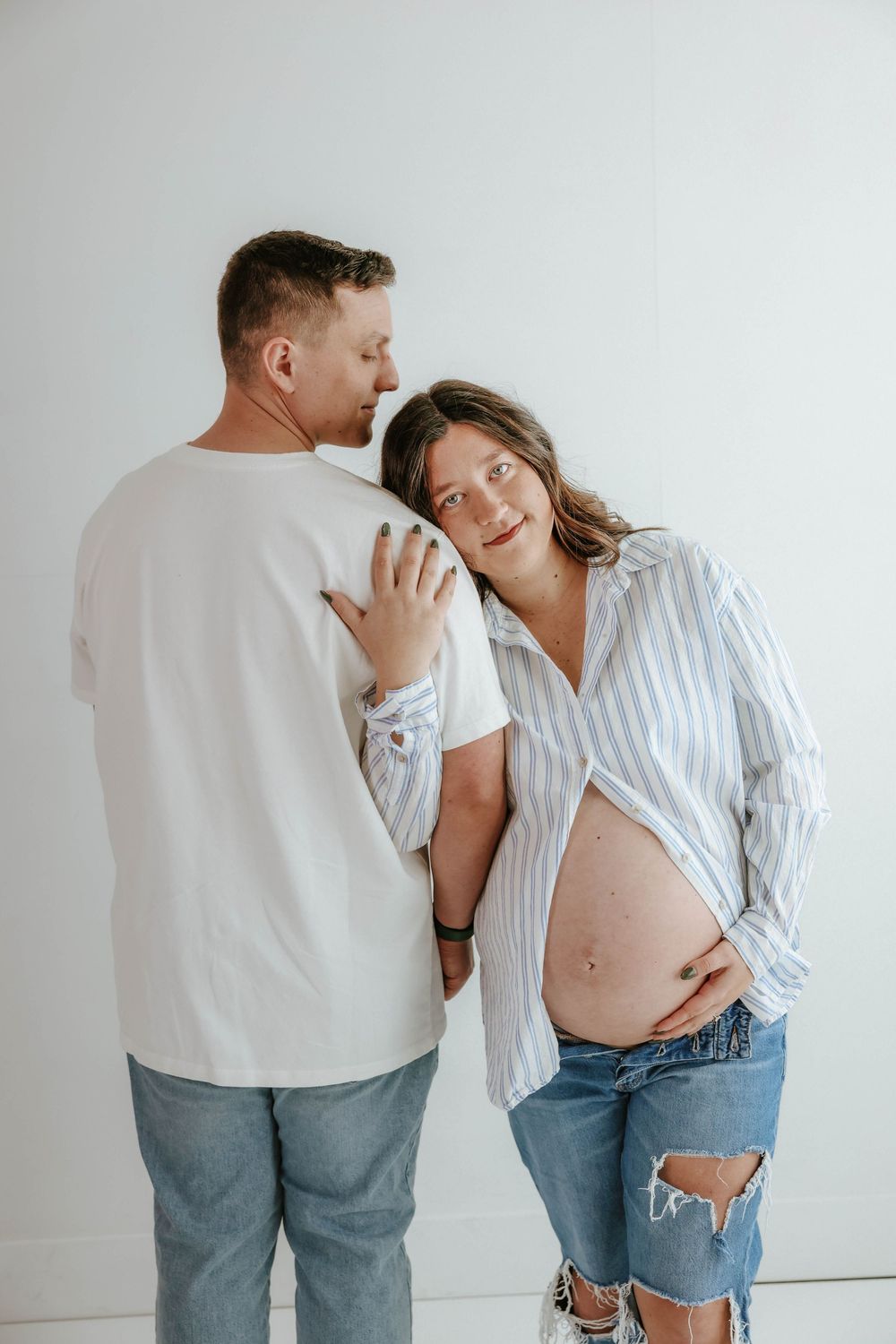 A couple shares an intimate moment against a white wall while showing off a growing baby bump in casual attire.