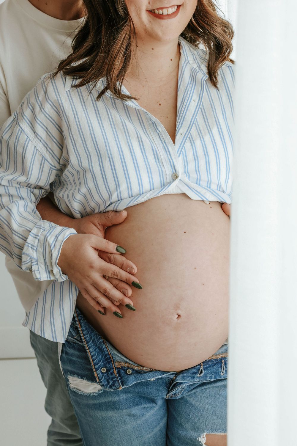 Pregnant belly being cradled while wearing a striped button-down shirt and denim jeans.