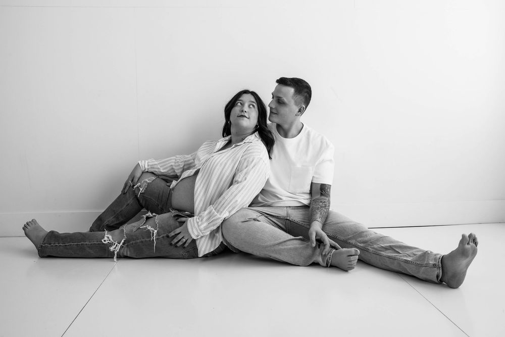 A couple in casual white outfits sits together on the floor in a black and white portrait against a plain wall.