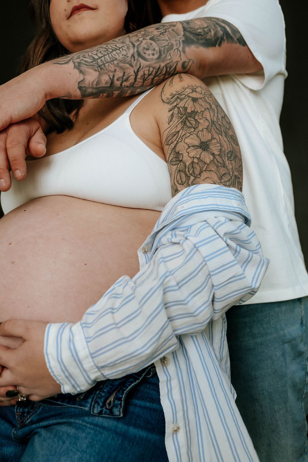Close-up of person with floral arm tattoos wearing white tank top, striped shirt and jeans in intimate maternity pose.