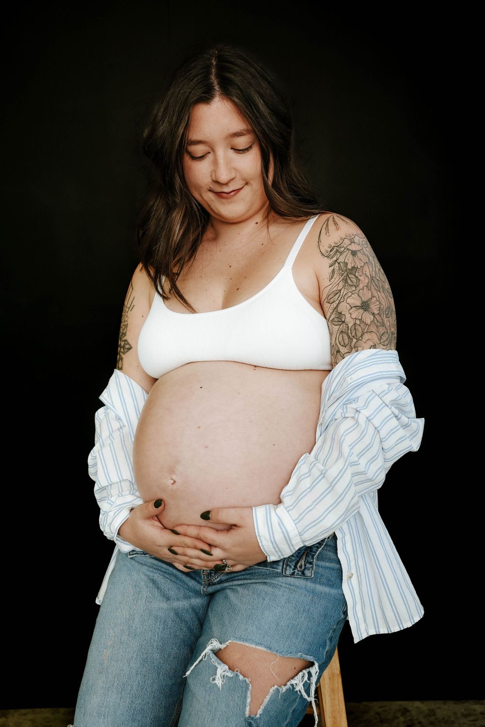 Expecting mother in white tank top and ripped jeans poses against dark background in maternity photoshoot.