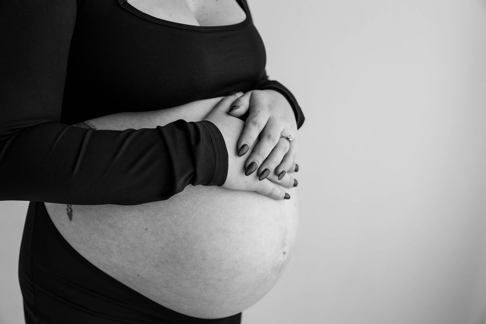 Black and white photo of hands gently resting on a pregnant belly, wearing dark clothing against light background.