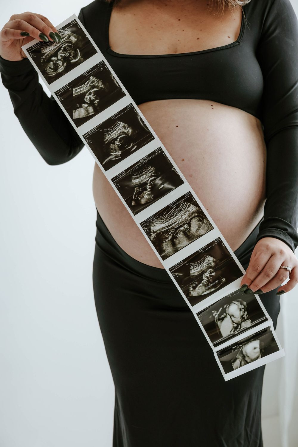 A pregnant woman in black clothing holds a strip of ultrasound photos across her baby bump against a white background.