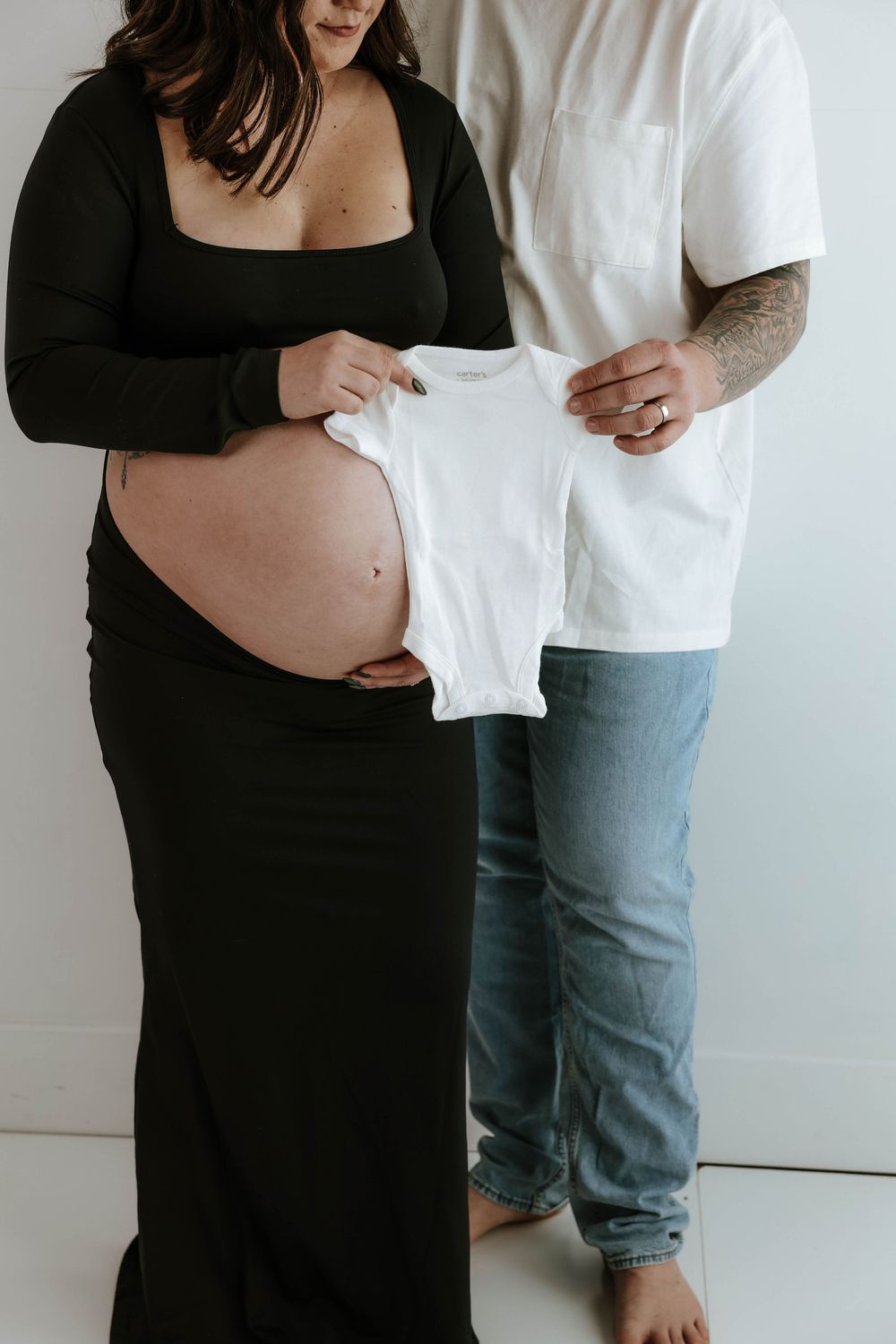 Pregnant woman in black dress holds white baby onesie with partner in jeans standing beside her against white wall.