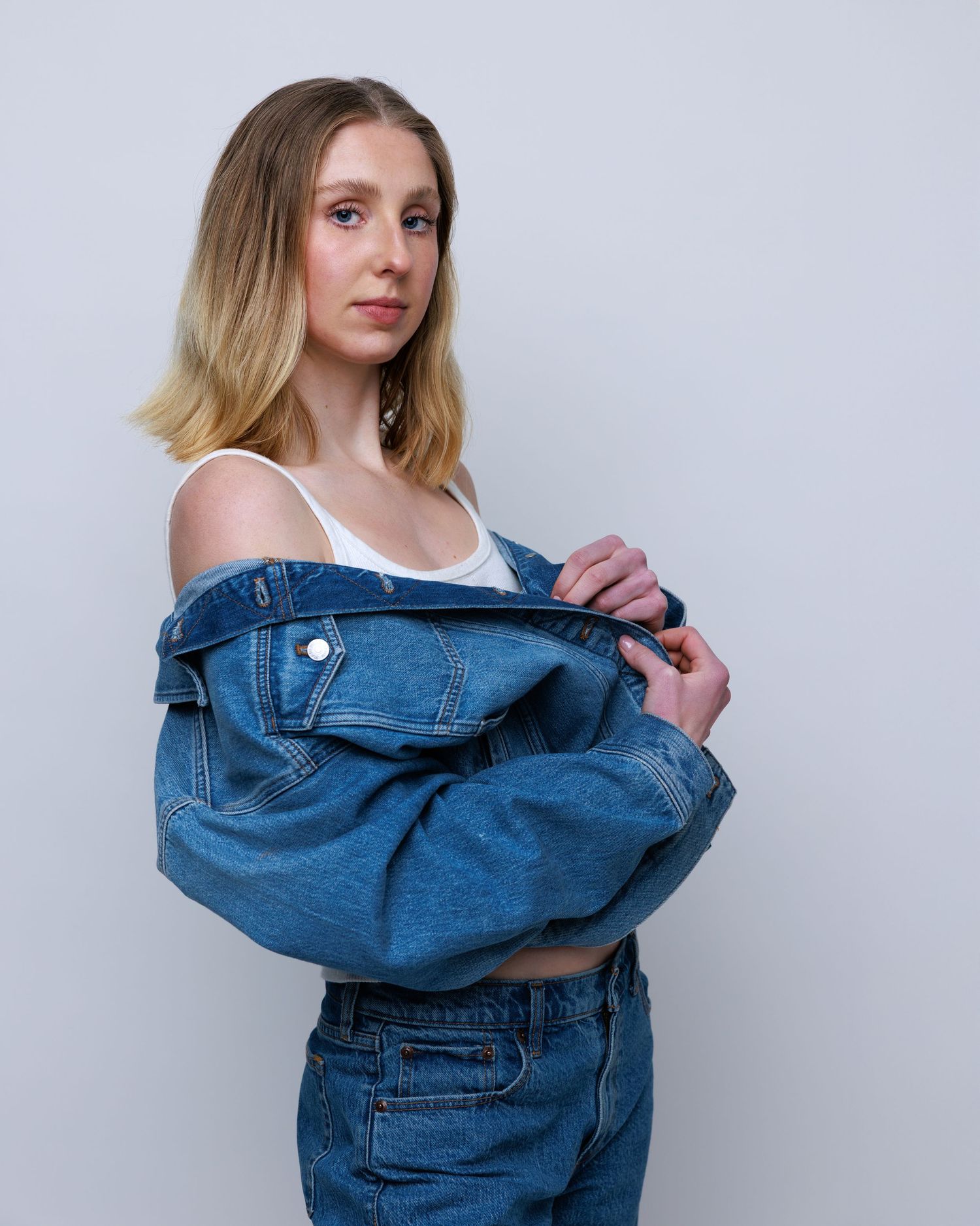 Casual portrait in white tank top and loose jeans against grey studio background.