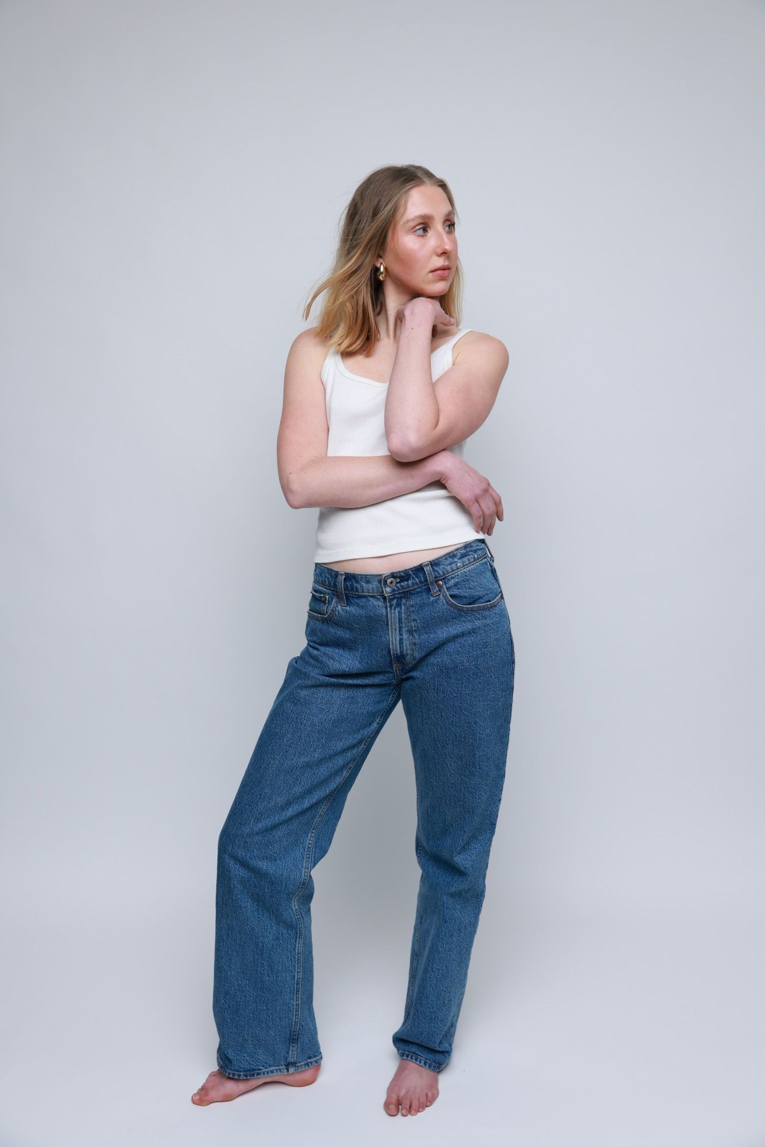 Casual portrait in white tank top and loose jeans against grey studio background.