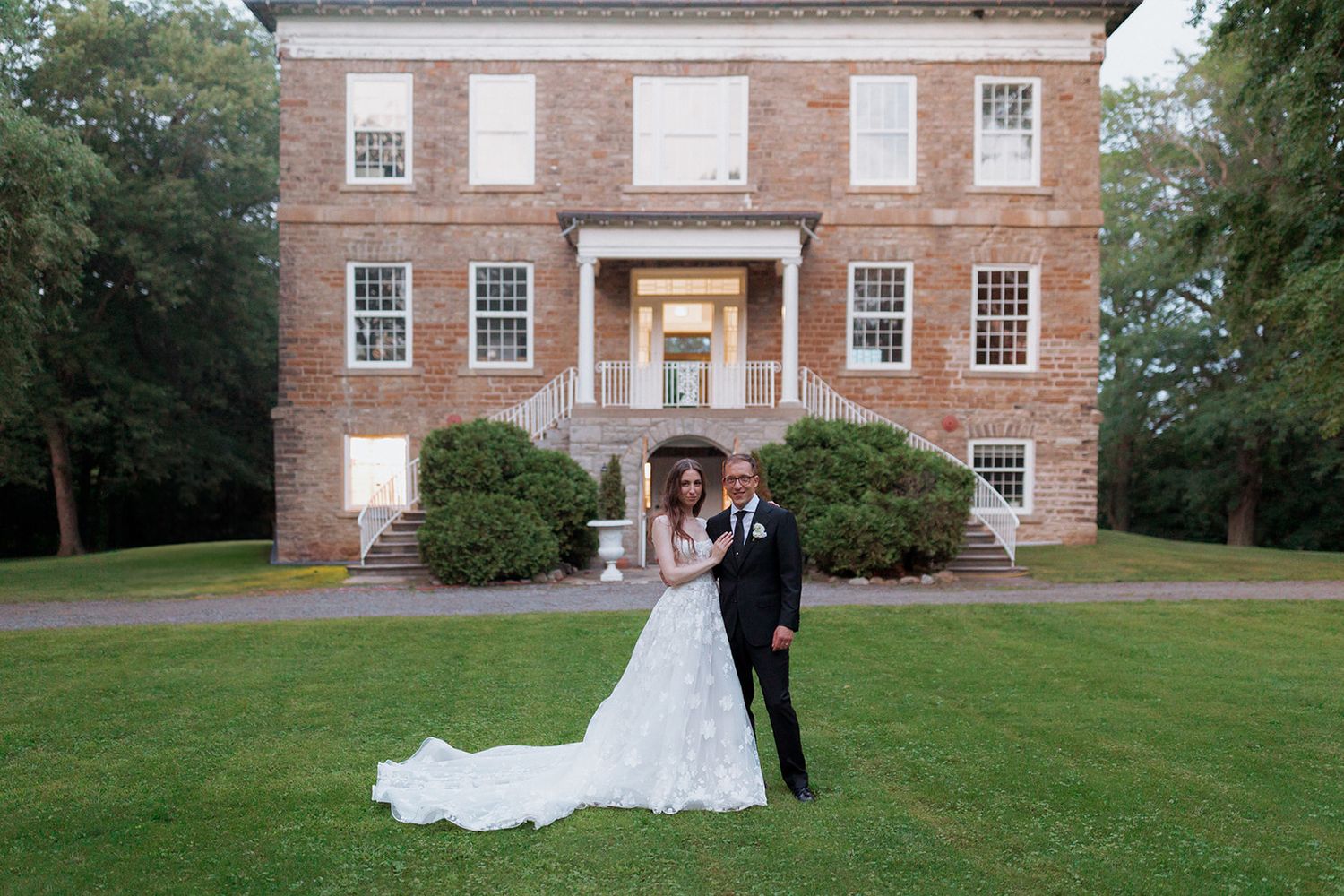 Wedding couple poses in front of historic brick mansion with landscaped grounds.