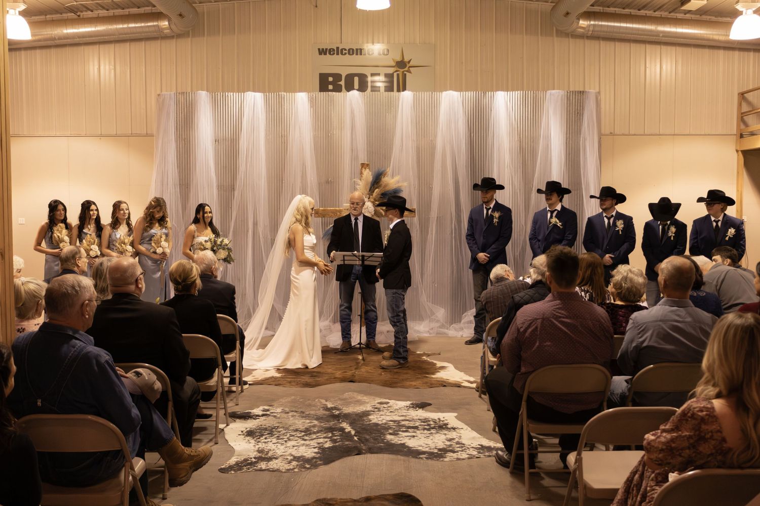 Indoor wedding ceremony with white draped backdrop and guests seated in rows at a reception venue.