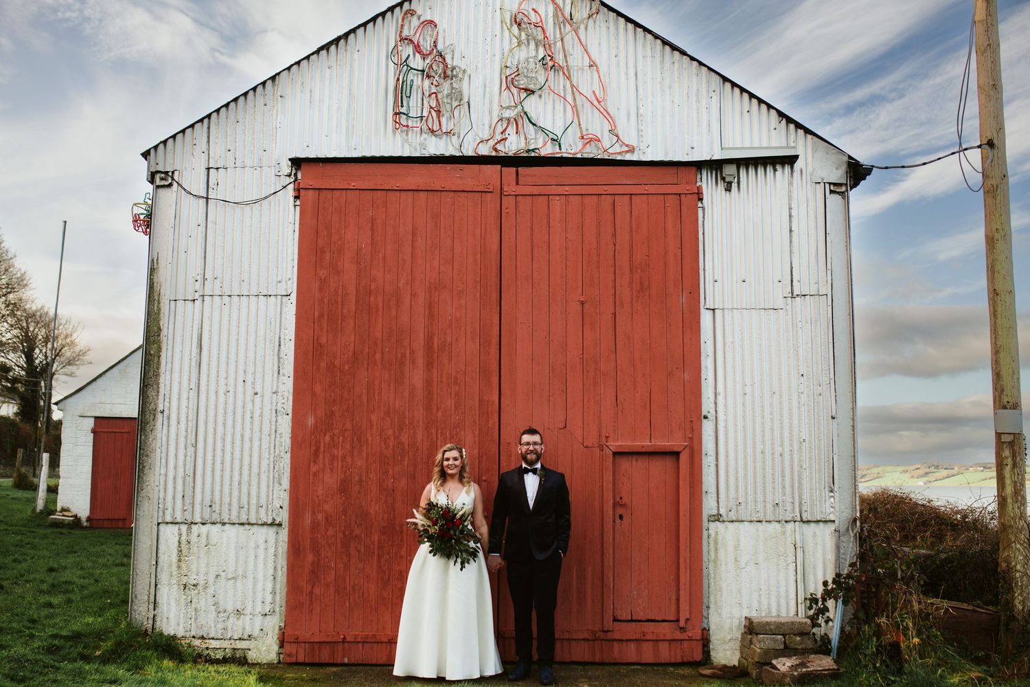 Rustic wedding couple stands in front of white barn with large red doors against cloudy rural sky.