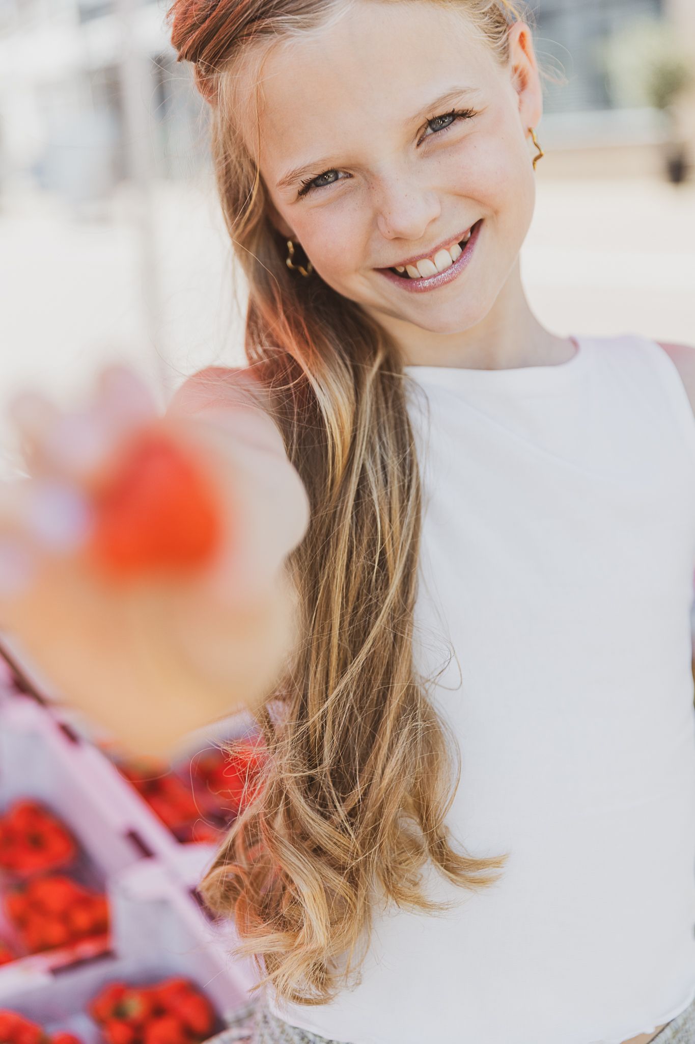 A person with long blonde wavy hair wearing a white top smiles warmly in a bright outdoor setting. Strawberrygirl. Kinderkleding fotografie