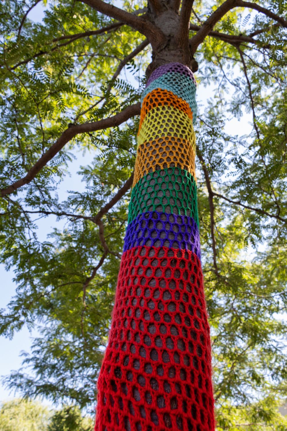 Rainbow colored yarn bomb covering a tree trunk in vibrant crochet patterns.