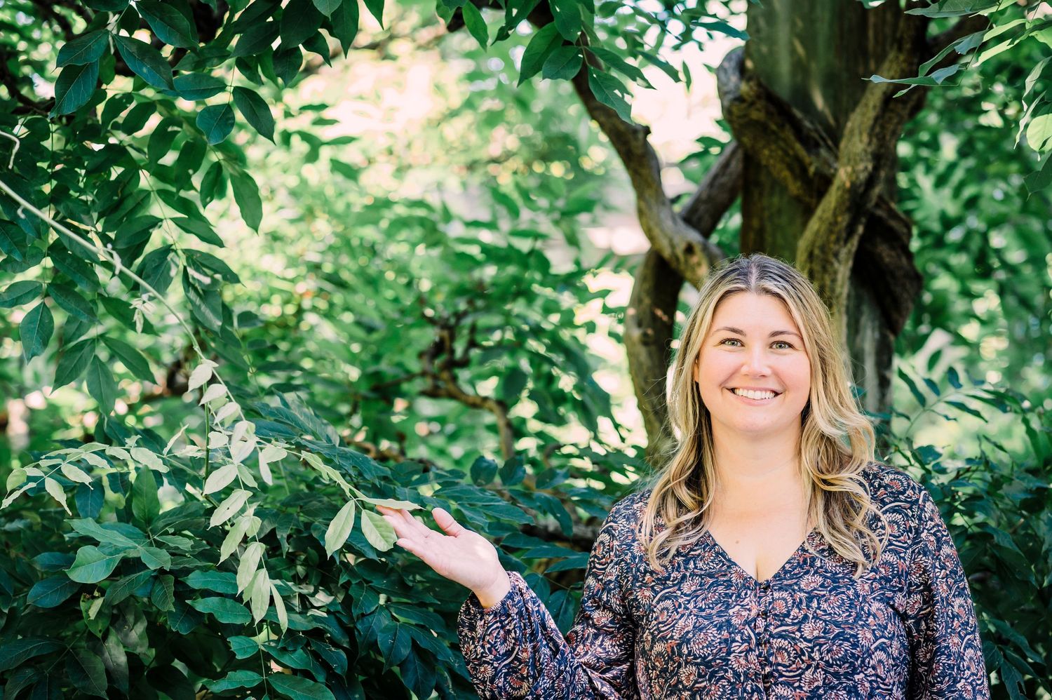 Person wearing blue floral top stands smiling in front of lush green foliage in outdoor garden setting.