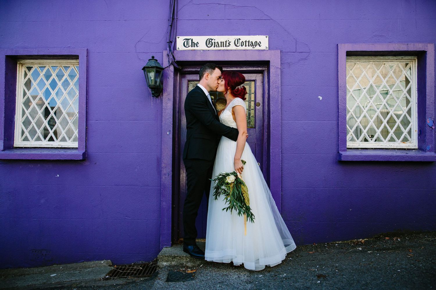 A couple shares a romantic moment outside a vibrant purple building with white latticed windows.