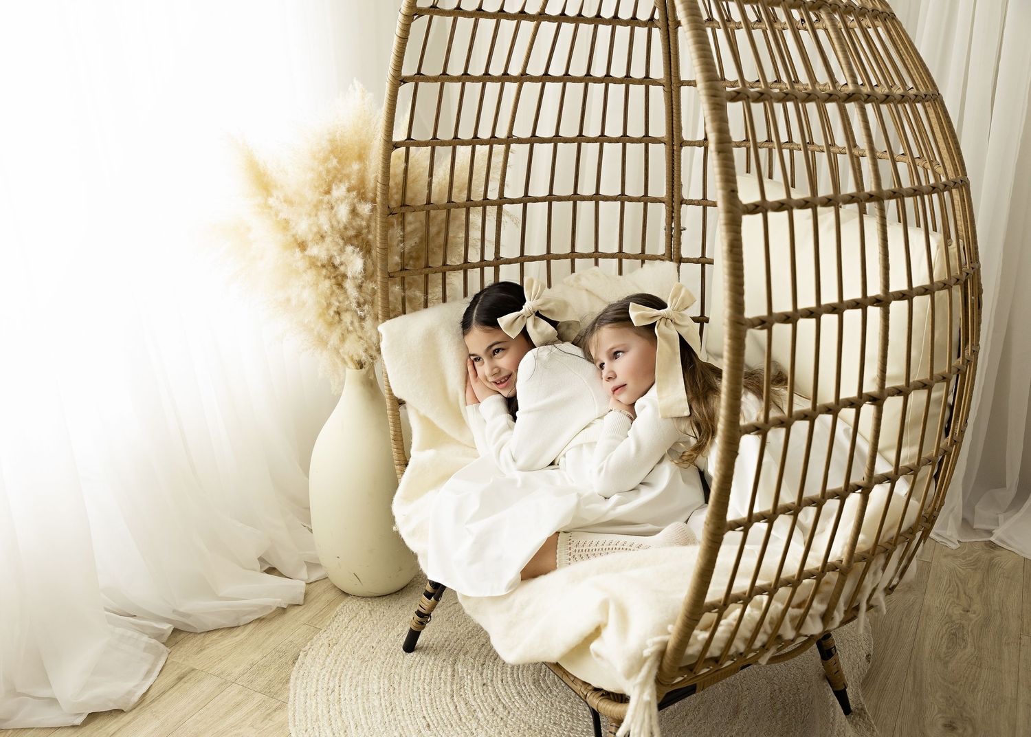Two children relax in a hanging rattan egg chair with white cushions and pampas grass decor.