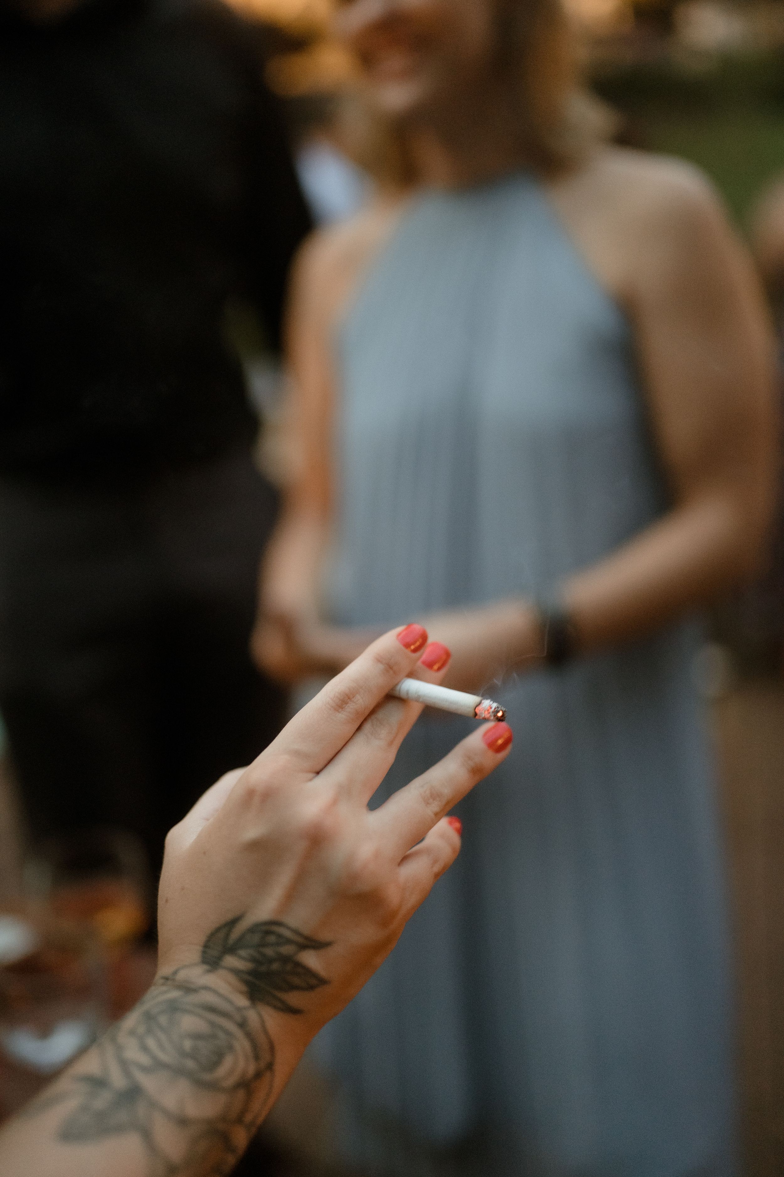 Hand holding a lit cigarette with visible rose tattoo artwork on the wrist.