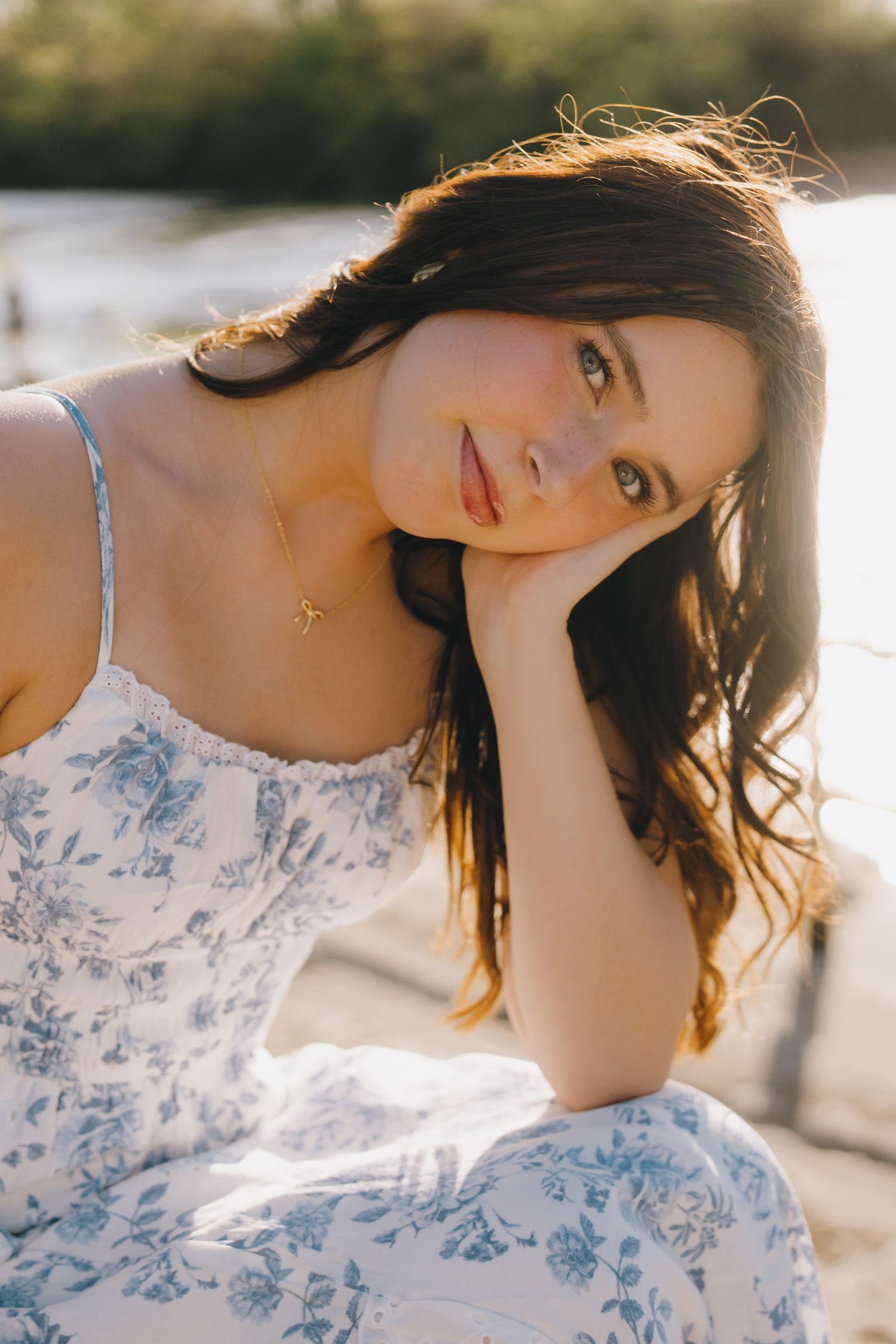 Sunlit portrait of a woman in a delicate blue and white floral dress with a gold necklace in warm evening light.