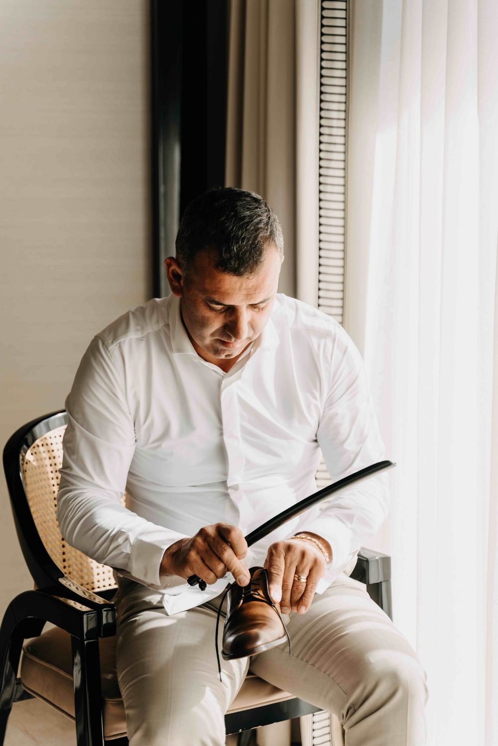 Man in crisp white shirt sits in rattan chair examining papers and documents in natural window light.