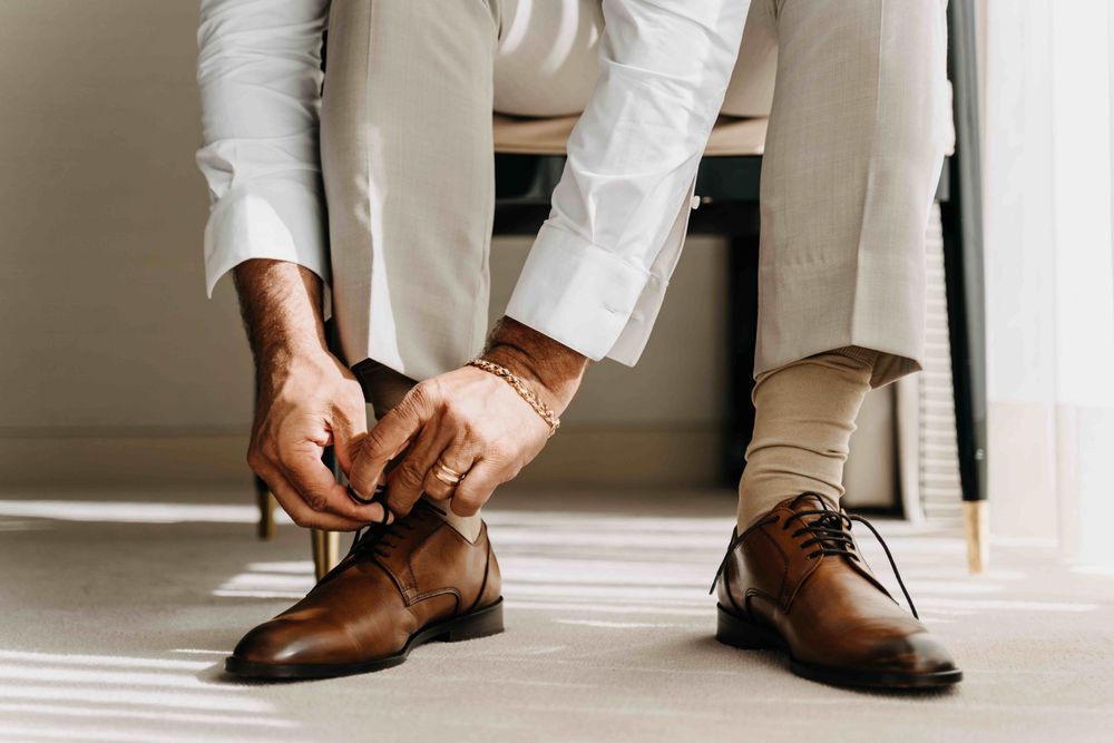 Close up of hands tying brown leather dress shoes with white pants and floor in background.