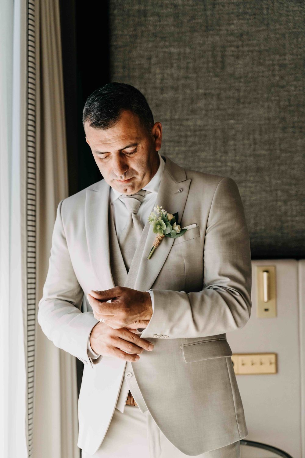 A groom in a light beige three-piece suit adjusts his cuff while wearing a white boutonniere.