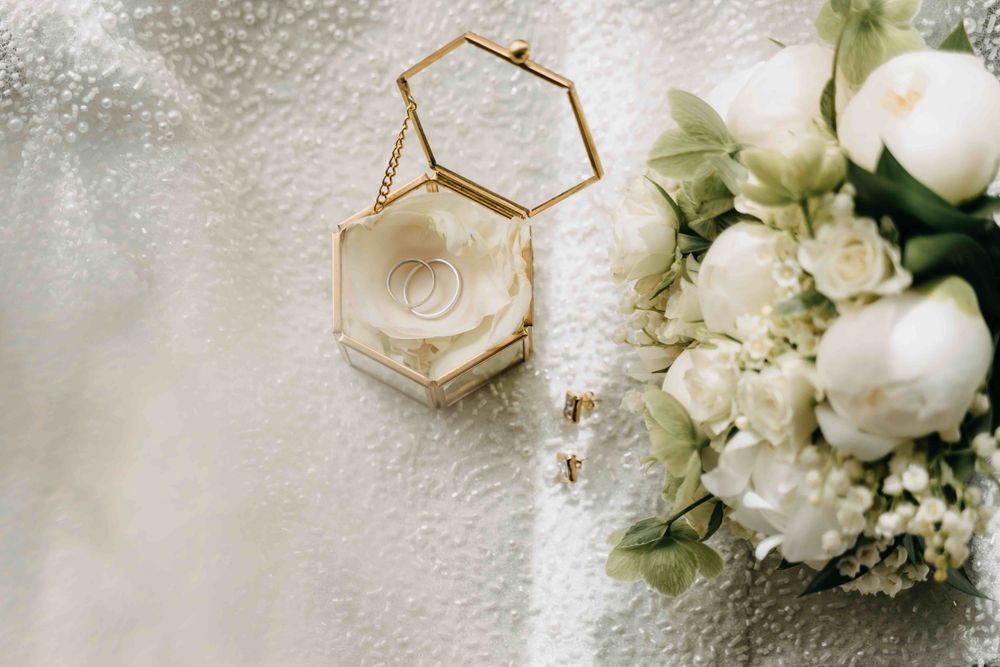 A hexagonal gold ring box rests beside white roses and floral arrangements on a textured white surface.