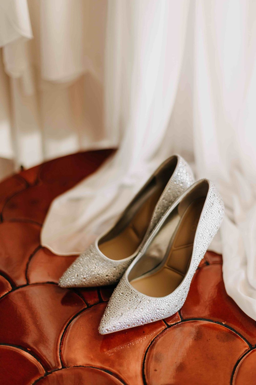 Sparkling crystal-encrusted white bridal shoes displayed on rich brown leather furniture.