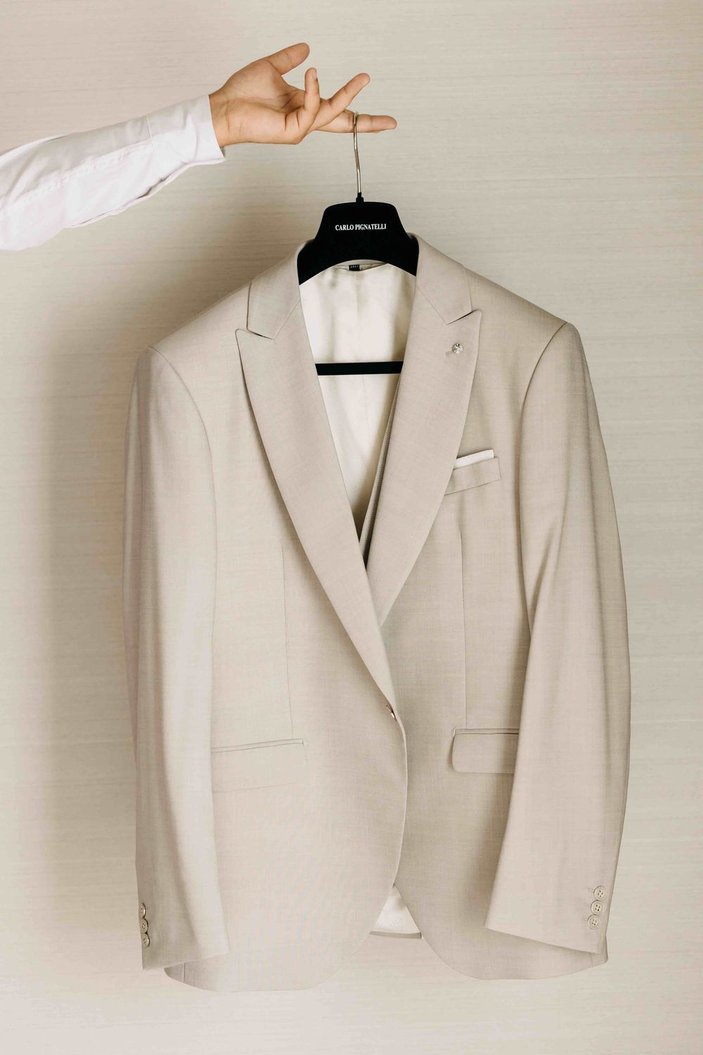 A crisp ivory white suit jacket hanging on a black hanger with a hand reaching to adjust it against a beige wall.