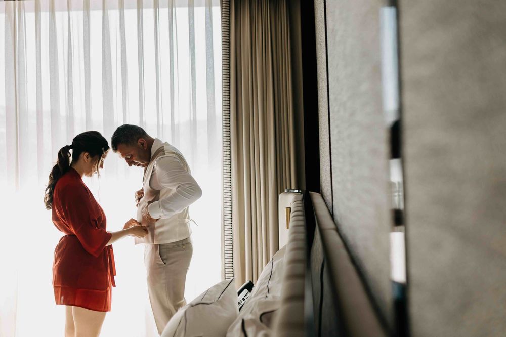 Two people sharing an intimate moment near a window with sheer curtains in a bright room.