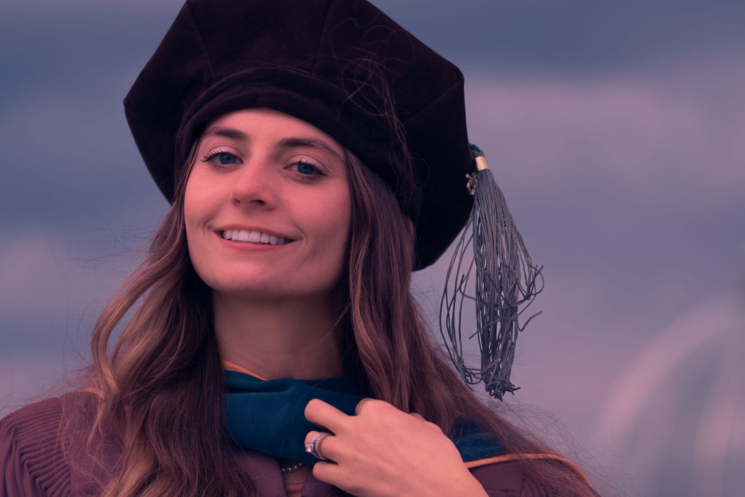 A young woman poses for grad photos from Wayne State University in black cap smiling during sunset against purple sky.