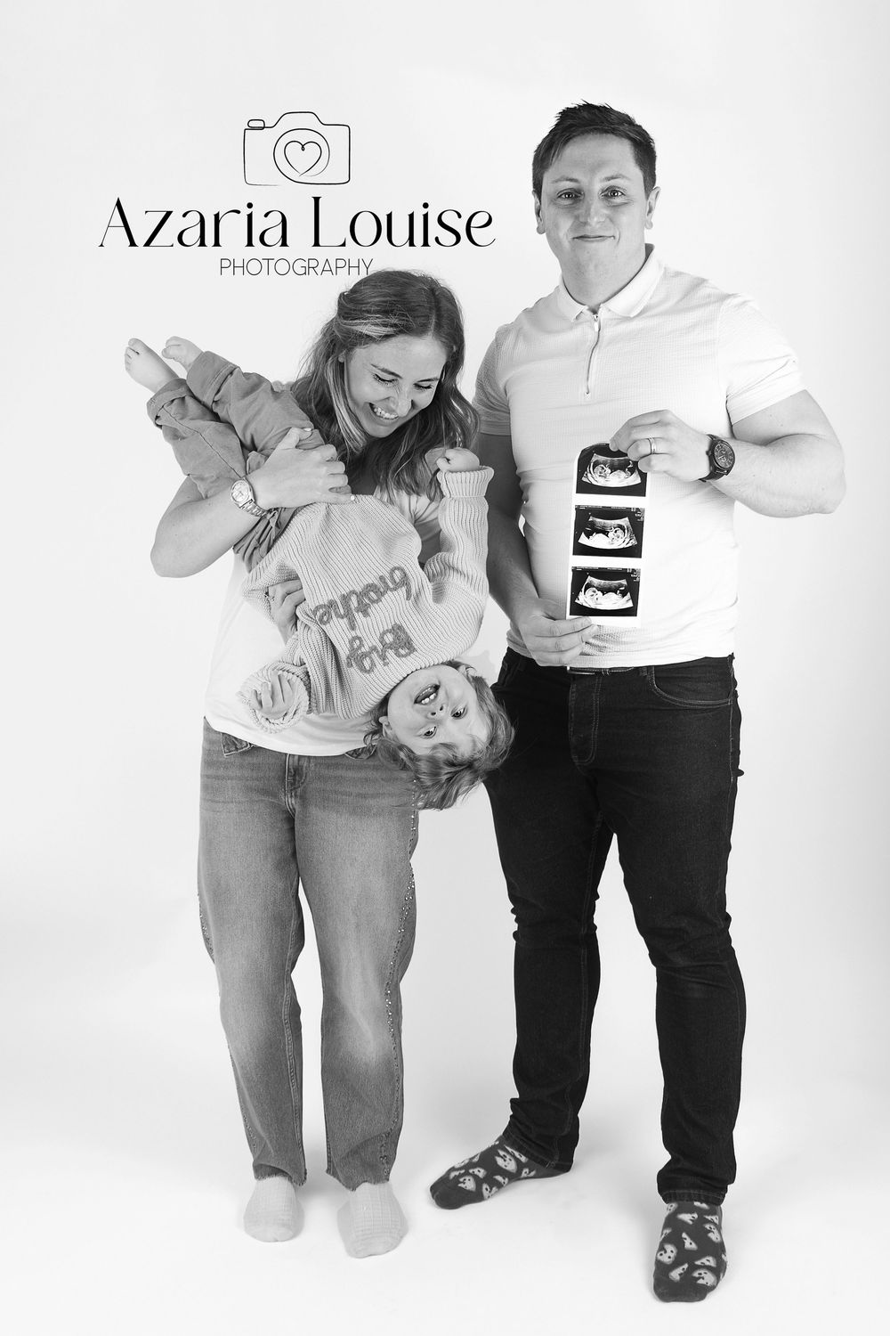 A black and white portrait of an expecting couple holding ultrasound photos and posing together in a studio setting.