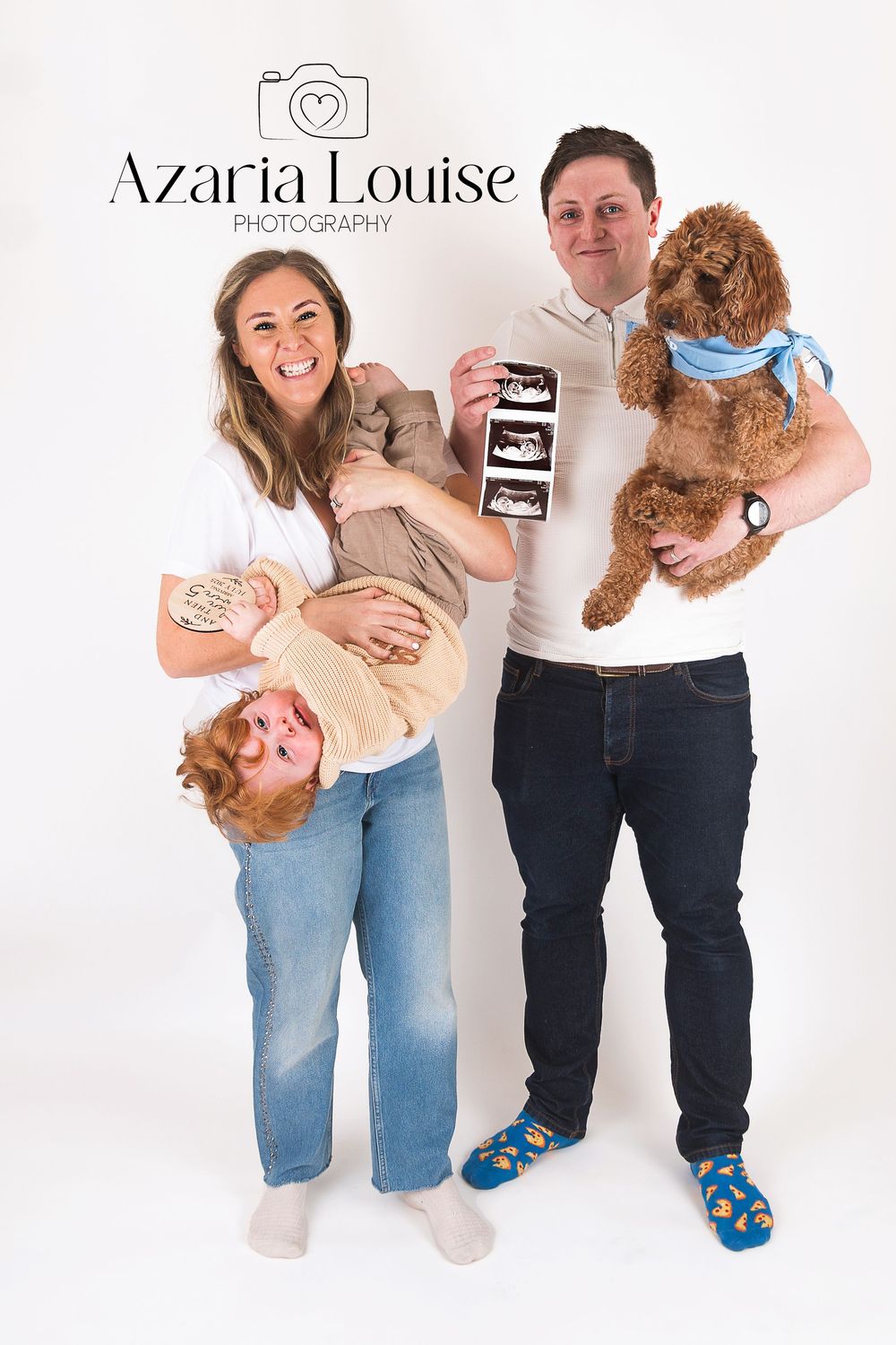 A young couple poses with their toddler and goldendoodle dog while holding ultrasound photos in a studio portrait.