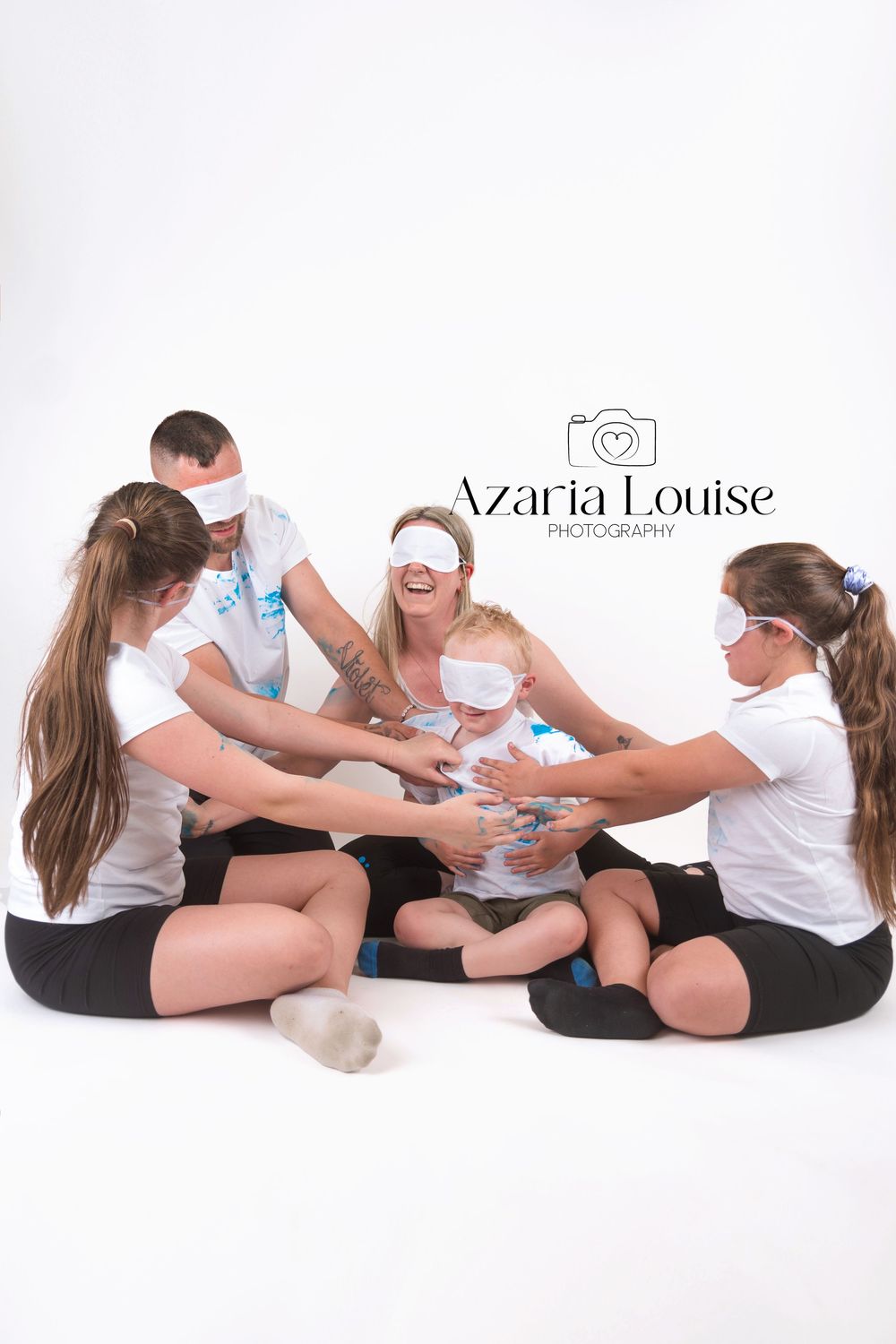 Family in white and black clothing sitting in circle on floor playing with small child against white backdrop.