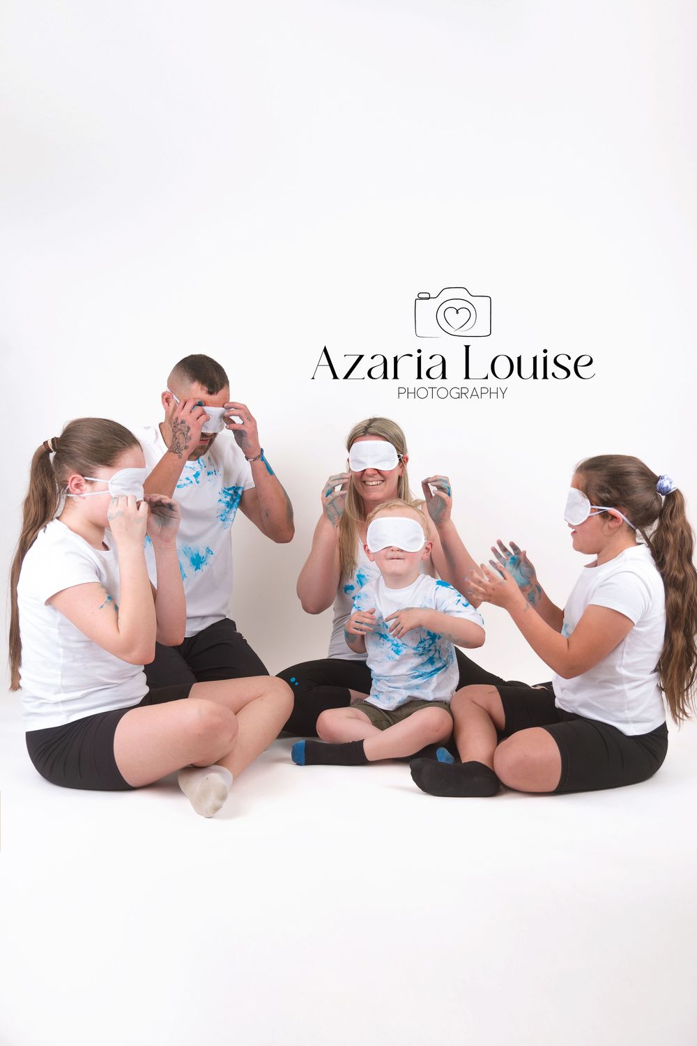 Group wearing white shirts and black shorts sit together having fun smashing cream pies in white studio setting.