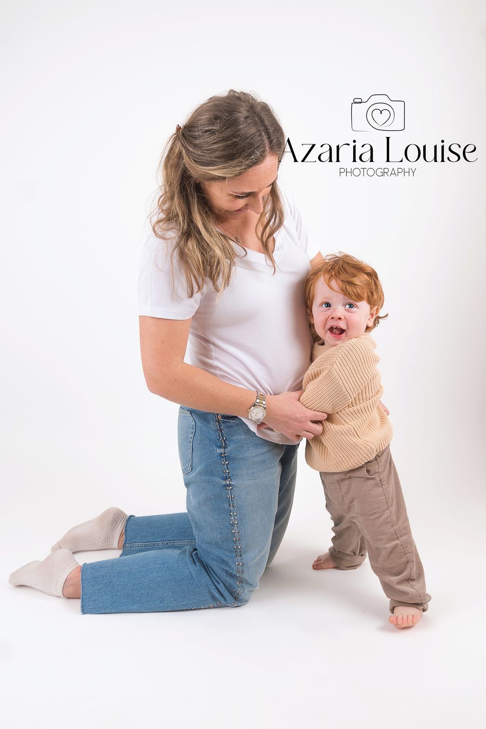 A parent in white shirt and jeans kneels beside a playful toddler wearing a beige sweater on a light background.