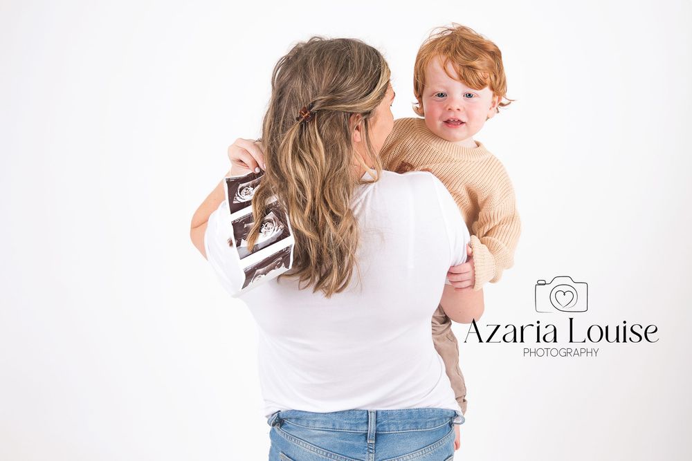 A parent in blue jeans and white shirt holds a small child wearing tan clothing against a white studio backdrop.