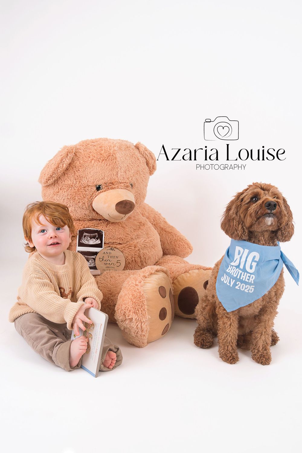 Baby in beige sweater sits next to large teddy bear and Goldendoodle dog wearing blue bandana in studio portrait.