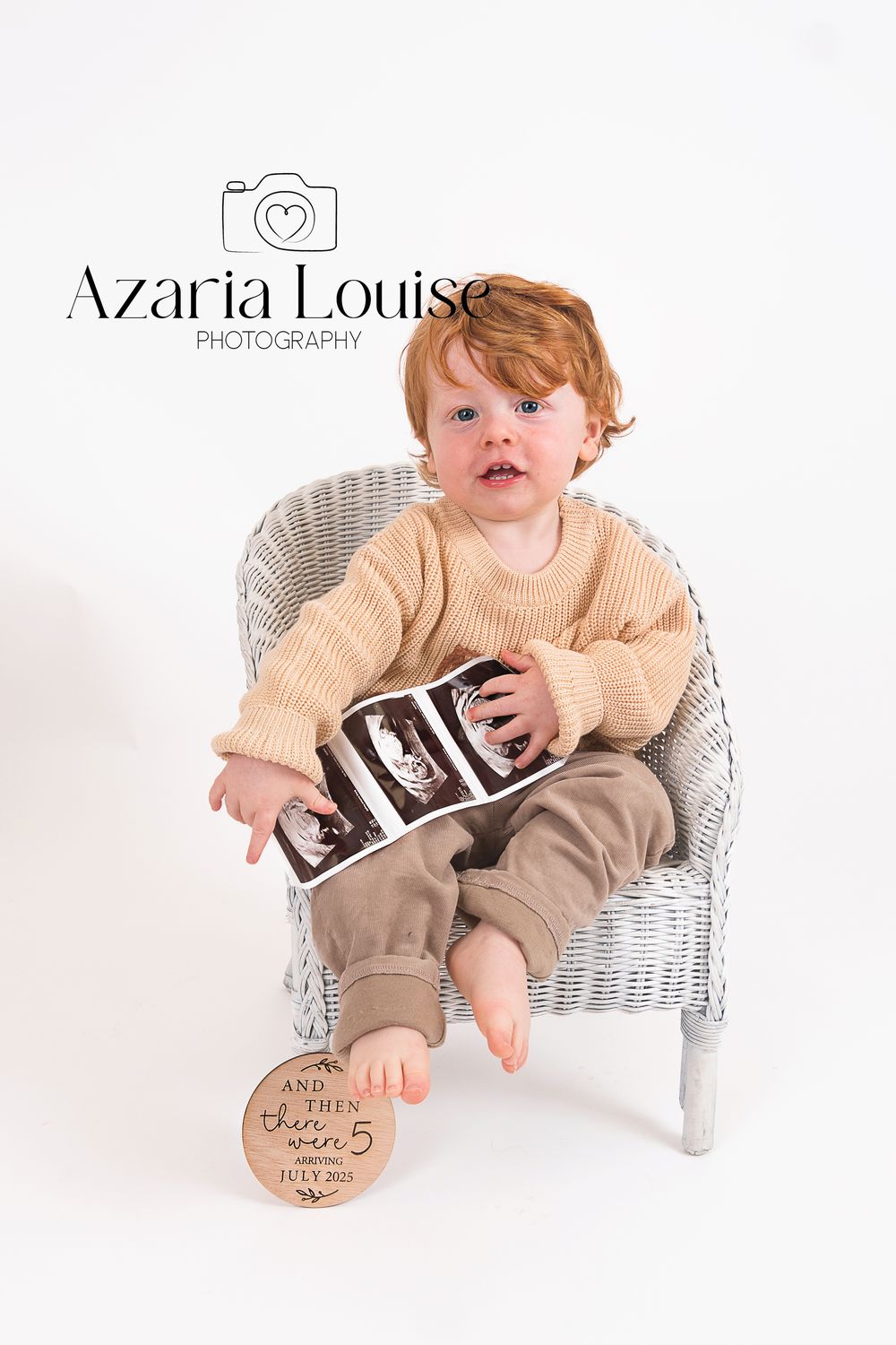 A young child in beige sweater sits in white wicker chair holding ultrasound photos during milestone photography session.