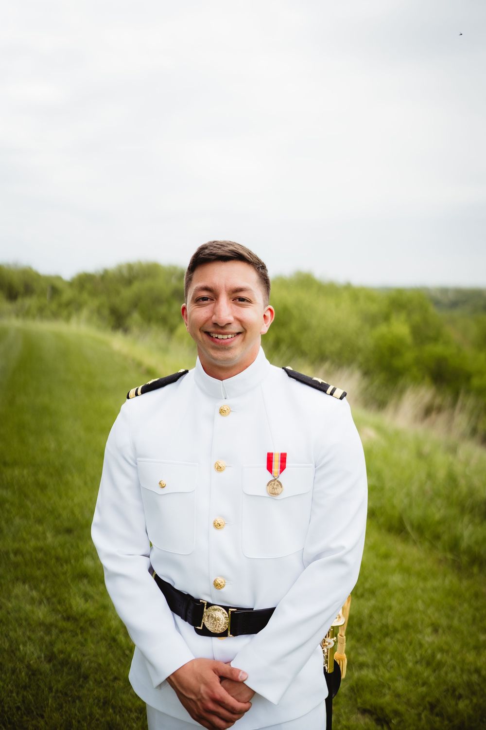 Military officer in white dress uniform with medal standing in grassy field under cloudy sky.
