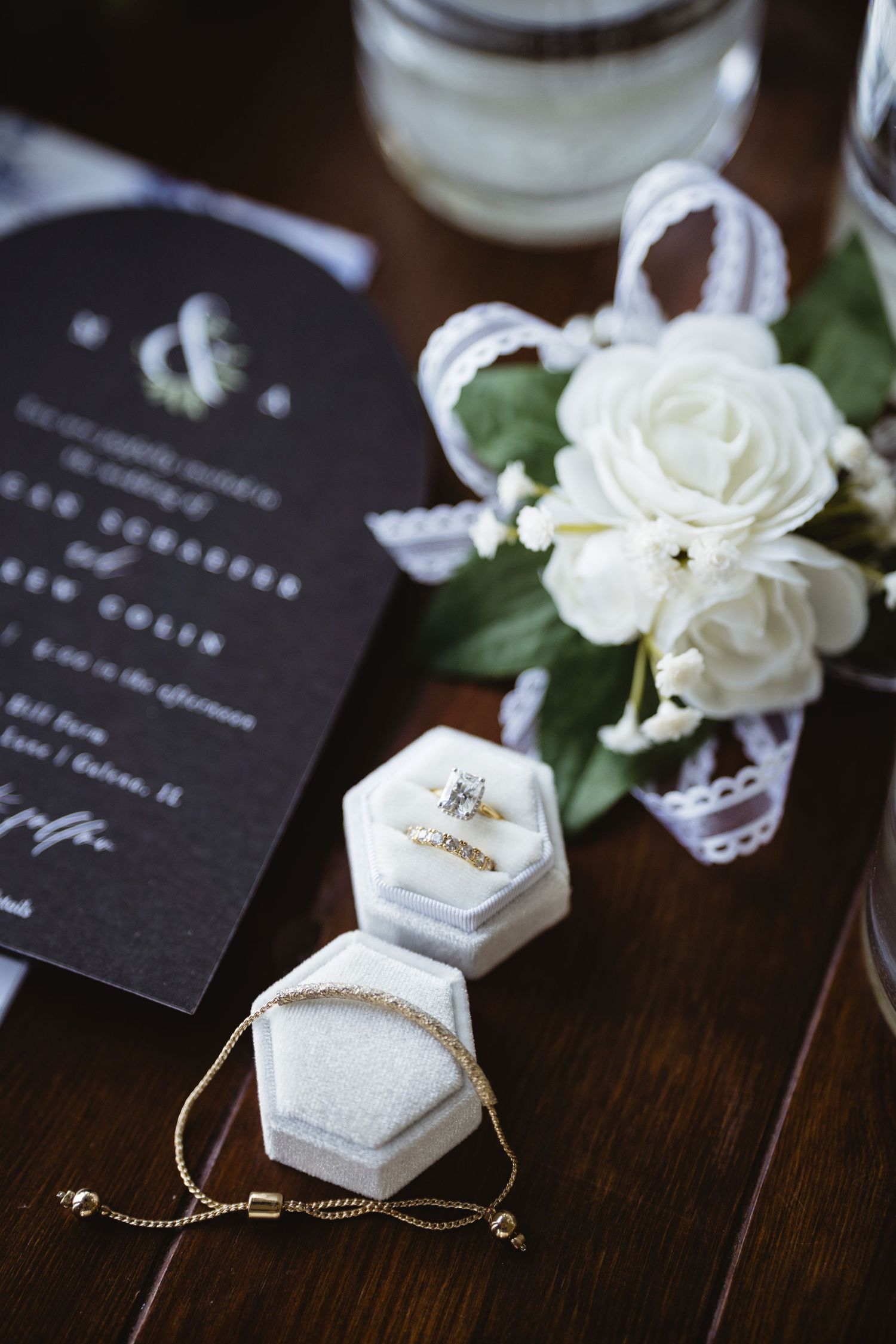 Wedding details featuring white roses, black invitation card, and marble ring boxes with delicate jewelry on dark wooden surface.