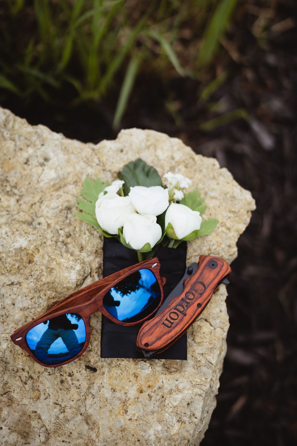 White roses, blue sunglasses, and a leather wallet arranged on a limestone rock with greenery in the background.