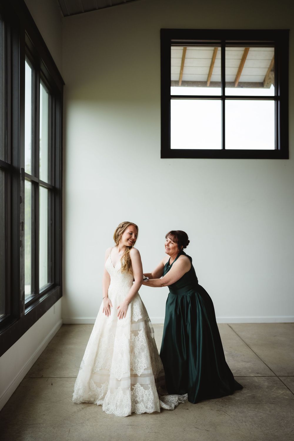 Two people share a candid moment near large windows in a bright room with one wearing an elegant lace gown.