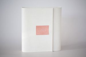 White paper notebook or journal with a small pink label or sticker on its cover against a light gray background.