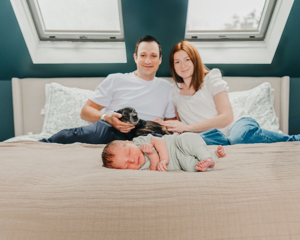 A couple relaxes together and cuddles on a white bed in matching white outfits while admiring their newborn baby