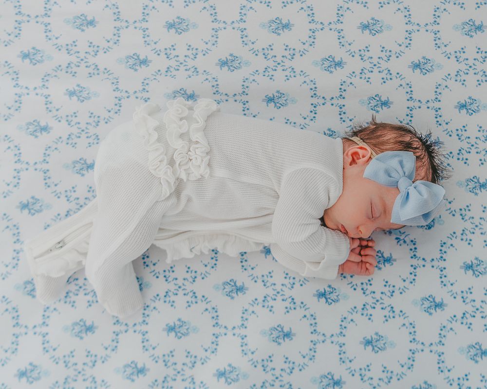 A baby sleeping peacefully on floral printed bedding in a white outfit and brown bow