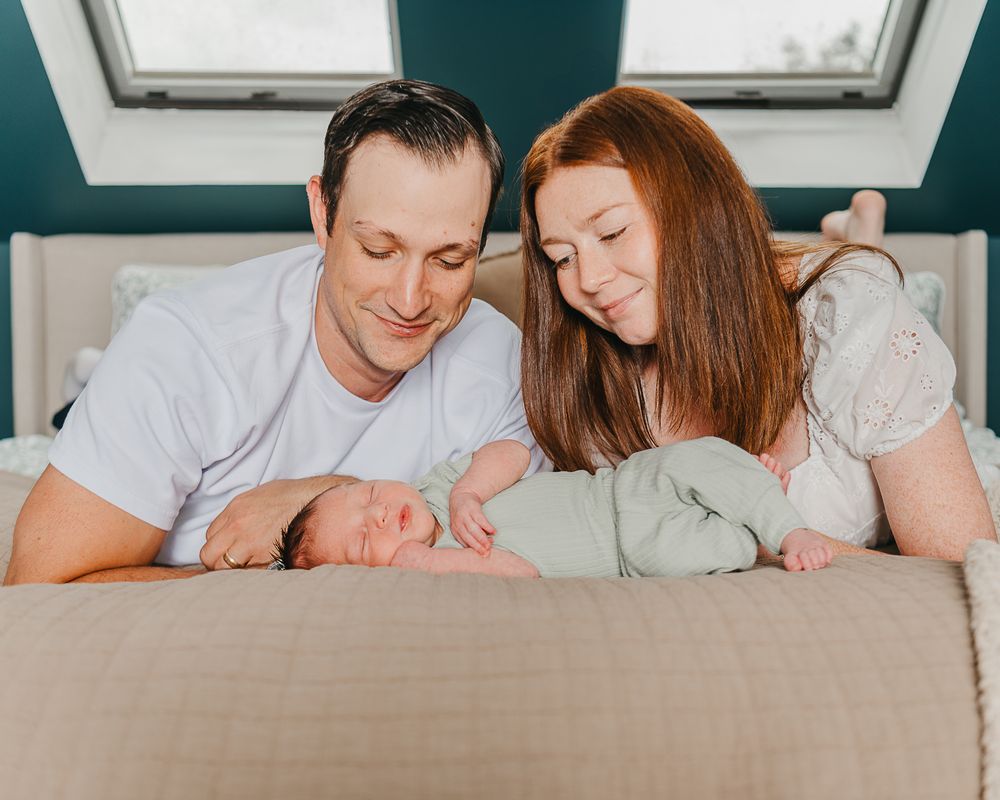 Parents lying on bed admiring their newborn baby in a cozy bedroom with teal walls and natural light