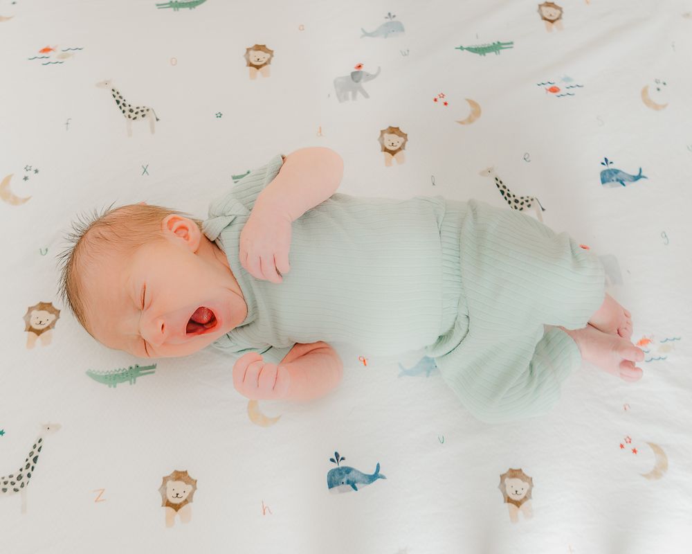 A sleeping baby wearing a mint green onesie lies on patterned bedding with whales and other whimsical designs